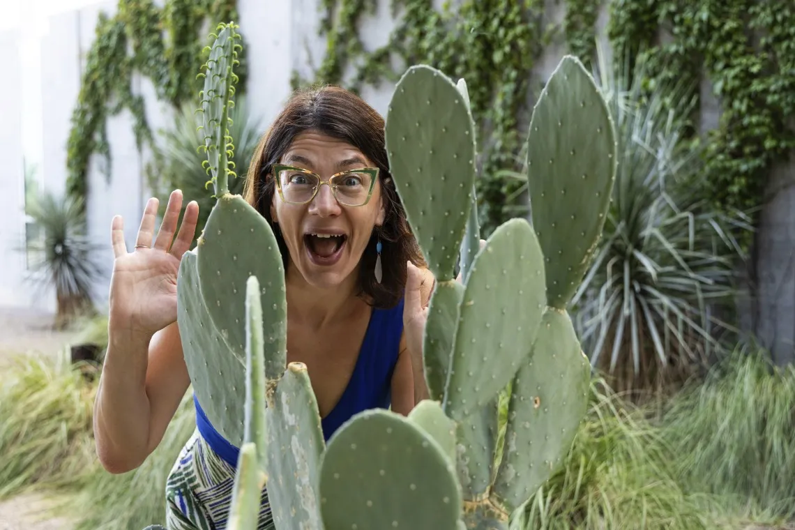 Image of Elise Gornish smiling and raising hand behind a cactus